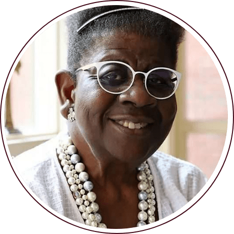 A person with glasses and a beaded necklace smiles while sitting by a window with natural light streaming in, exuding the calm resolve reminiscent of civil rights pioneers. They have short, curled hair and are wearing a white shirt.