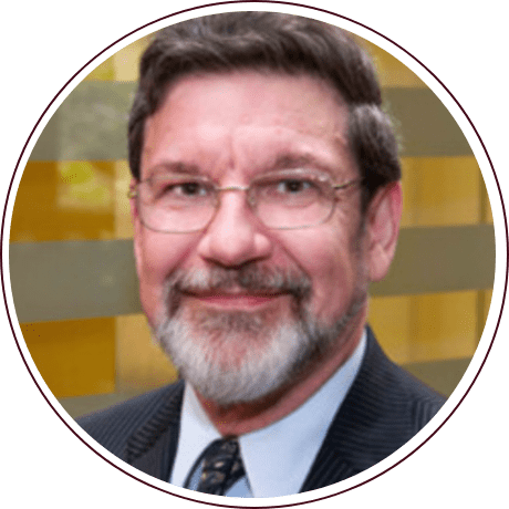 A man with glasses and a beard is smiling at the camera, embodying the spirit of advocacy seen at the Fred Gray Institute. He is wearing a dark suit and tie, standing against a backdrop of horizontal gold and brown stripes.