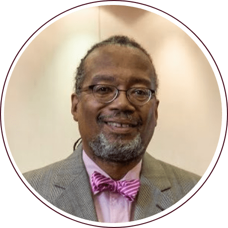 A man with glasses wearing a gray suit and pink bow tie stands in front of a neutral background, channeling the elegance of civil rights advocate Fred Gray, as he looks at the camera with a slight smile.