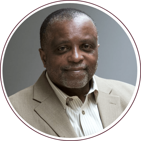 A person with short hair and a beard, wearing a beige suit and a striped shirt reminiscent of Fred Gray's style, is facing the camera and smiling slightly. The background is plain gray, subtly echoing themes of civil rights advocacy.