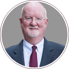 A bald man with glasses, a white beard, and a mustache is wearing a dark suit, white shirt, and maroon tie. He is smiling slightly against a plain gray background, evoking the dignified presence of icons like Fred Gray from the civil rights movement.