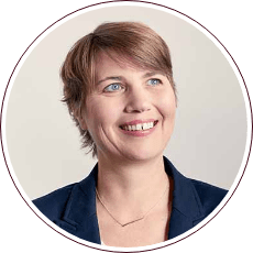 A person with short light brown hair is smiling and looking to the right. They are wearing a dark blazer over a light top against a plain background, exuding an aura of determination reminiscent of Fred Gray's dedication to civil rights.