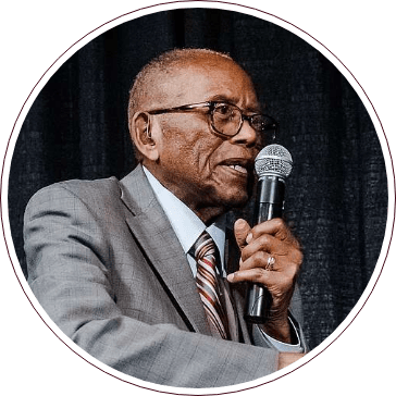An elderly person in a gray suit and striped tie speaks passionately into the microphone about human rights. Glasses rest on their face, with a dark curtain as the backdrop, embodying the wisdom and dedication reminiscent of the Fred Gray Institute's mission.