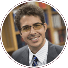 A man with curly hair and glasses is smiling, exuding warmth. He is wearing a suit and tie, standing confidently in front of a blurred view of bookshelves that whisper stories of justice from the Fred Gray Institute for human rights.