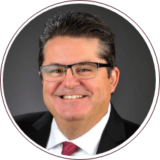 Man with short dark hair, wearing glasses and a suit with a red tie, smiling against a dark background.