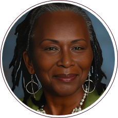 A person with long hair and hoop earrings is smiling in a portrait photograph. They are wearing a green top and a pearl necklace, with a neutral blue-gray background.