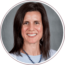 A woman with shoulder-length brown hair is smiling. She is wearing a white top and a blue beaded necklace. The background is plain gray.