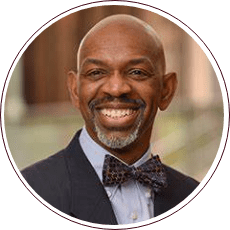 A person wearing a dark suit, light blue shirt, and patterned bow tie is smiling. The background is blurred, showing shades of brown and green.