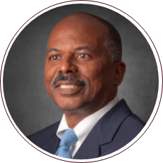 A man in a suit and tie smiles against a gray textured background.
