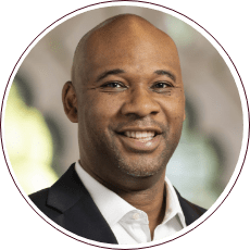 A person with a shaved head is smiling while wearing a suit and white shirt. The background is softly blurred, highlighting the individual's face and expression.