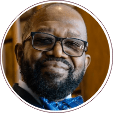 A man with a beard and glasses is smiling slightly. He is wearing a blue bow tie and a suit. The background is blurred, showing warm-toned wooden elements.