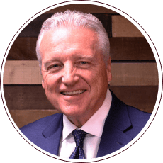 A smiling man with white hair is wearing a suit and tie, standing against a background of wooden panels.