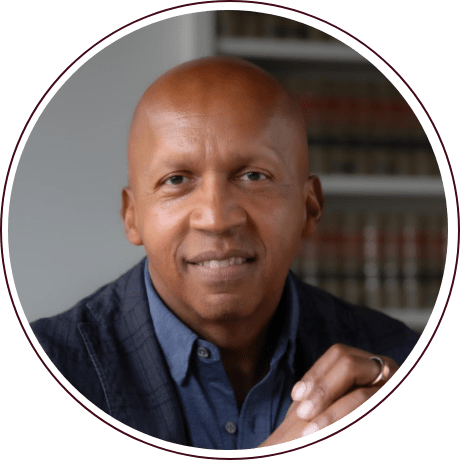 A person with a shaved head and a light smile is wearing a navy blue shirt and a dark blazer. They are sitting in front of a blurred background of bookshelves and resting their hand on a table.