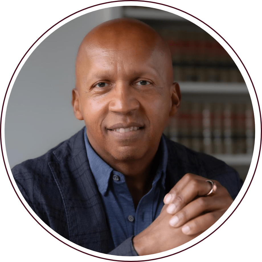 Bald person wearing a dark blazer and blue shirt, smiling gently, seated with hands clasped. Blurred background with bookshelves.