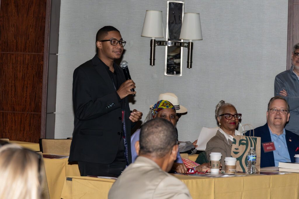 A person stands holding a microphone, speaking to an audience at a conference. Others sit at tables with yellow tablecloths, listening. A Starbucks cup, papers, and a gift bag are visible on the table. Wall lights are on the gray background.