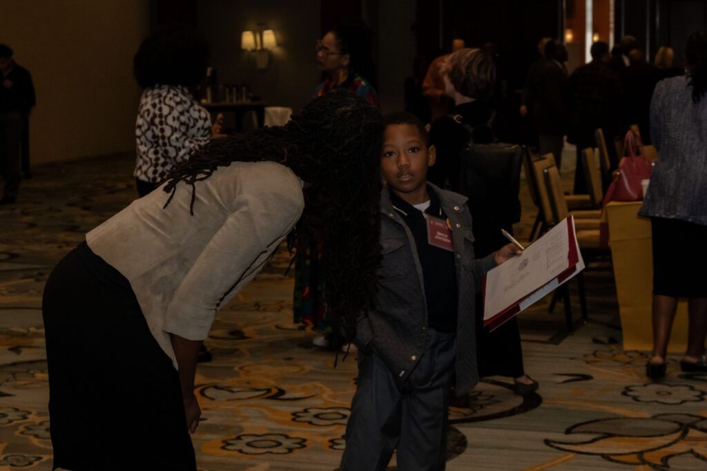 A woman with long hair leans down to speak to a young boy holding a clipboard in a conference room filled with people. The scene has a carpeted floor with a patterned design and chairs arranged in rows.