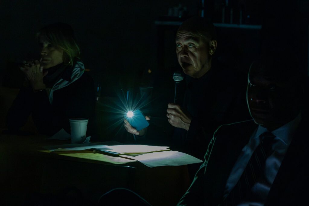 A group of people in a dimly lit room sit around a table. One person holds a microphone in one hand and a phone emitting a bright light in the other. Papers and a cup are on the table in front of them.