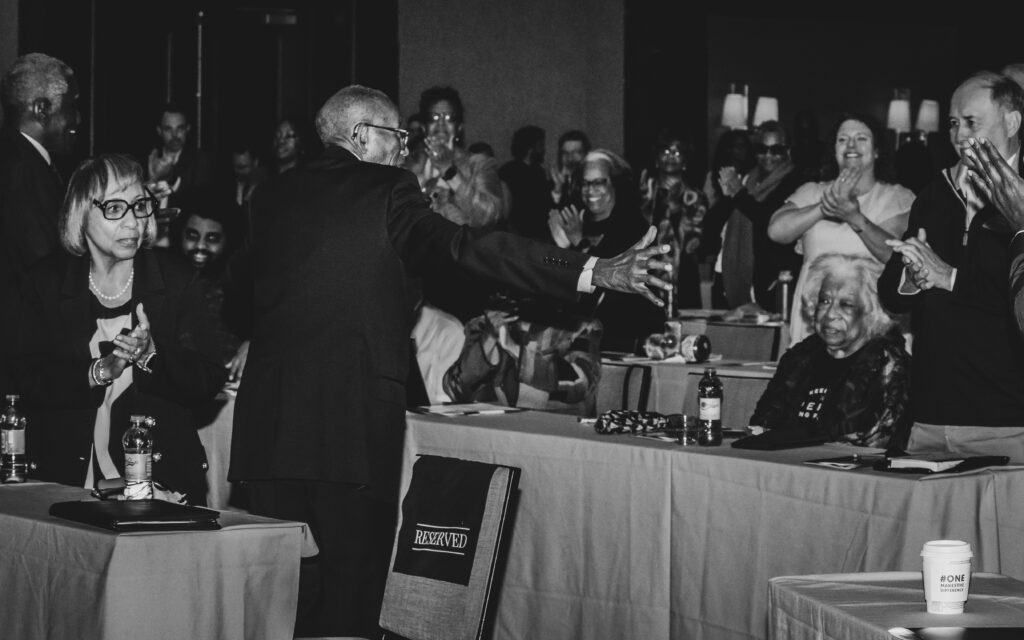 A black-and-white photo of a man facing and gesturing towards an applauding audience in a conference room. Seated and standing people are visible, some smiling and clapping. Tables with items on them are in the foreground.