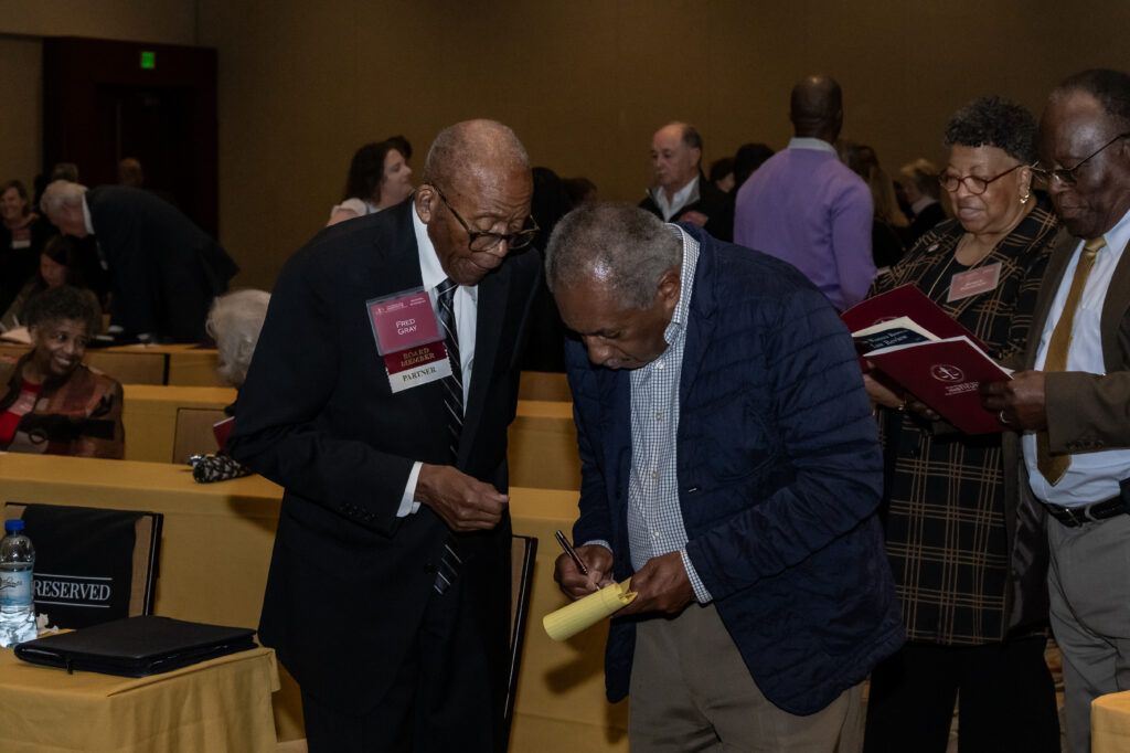 Two elderly men stand in conversation at an event; one wears a suit with name tags and the other signs a paper on a yellow notepad. People in the background are seated or standing, some holding booklets. The setting appears to be a formal gathering or conference.