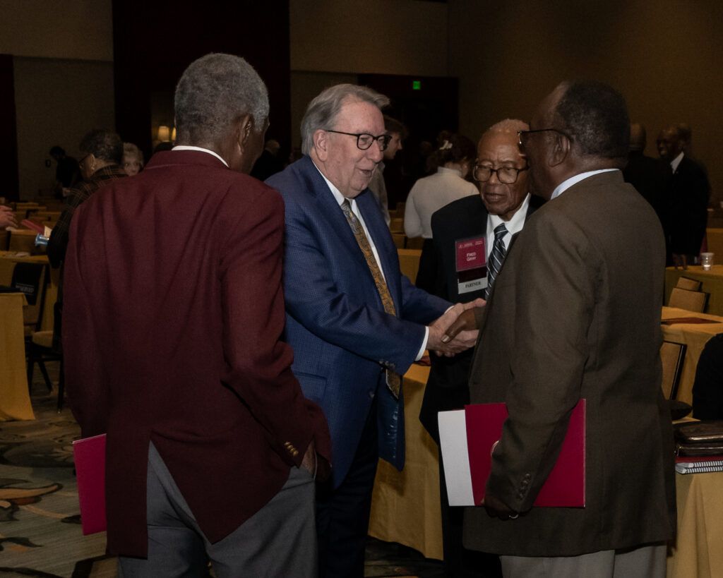 Four men are conversing in a dimly lit conference room. One man, wearing a blue suit, is shaking hands with another man in black attire. Two other men in suits, one holding a red folder, are observing the handshake.