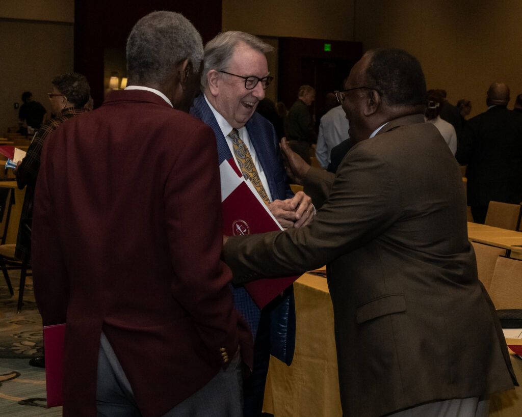 Three men in suits are standing and talking in a conference room. Two of them are holding red folders. The background shows several tables and chairs, and other people are visible in the dimly lit setting.