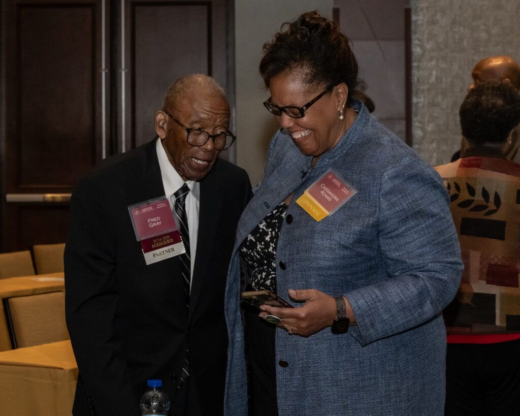 Two people are standing and smiling in a conference room. The man on the left is wearing a suit and tie with a name badge. The woman on the right is wearing glasses, a blue jacket, and holding a phone. Conference materials are visible in the background.