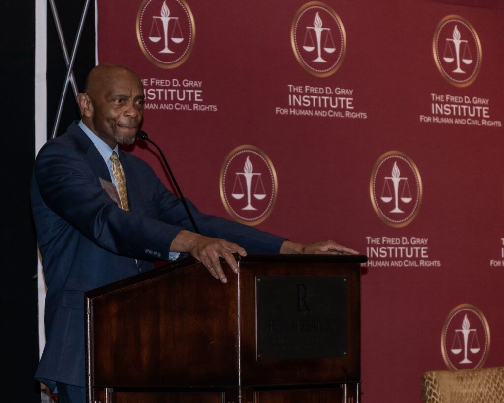 A person stands at a podium speaking into a microphone. The backdrop has logos and text that read "The Fred D. Gray Institute for Human and Civil Rights." The event appears to be held in a formal setting.