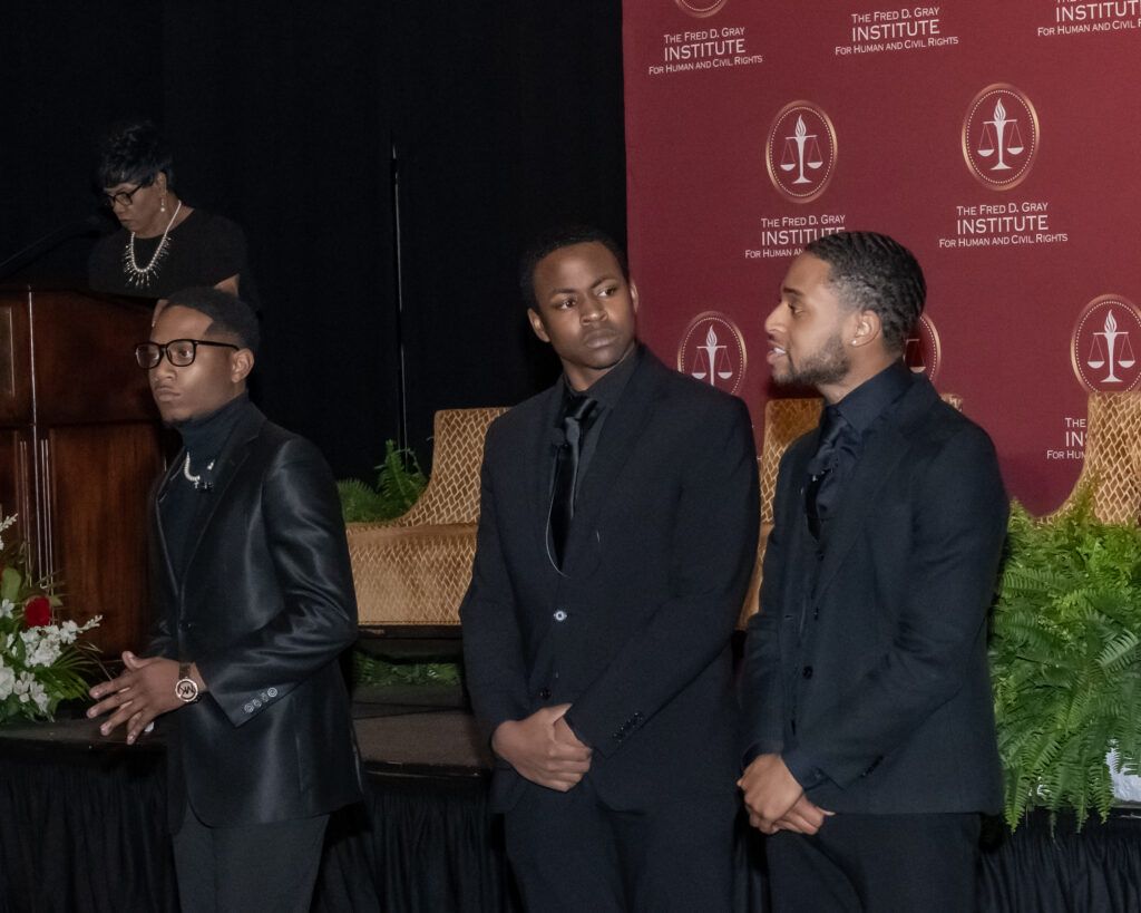 Three men in suits stand on a stage near a podium, with a woman speaking in the background. A red banner with "FRED D. GRAY INSTITUTE for HUMAN and CIVIL RIGHTS" is partially visible behind them.