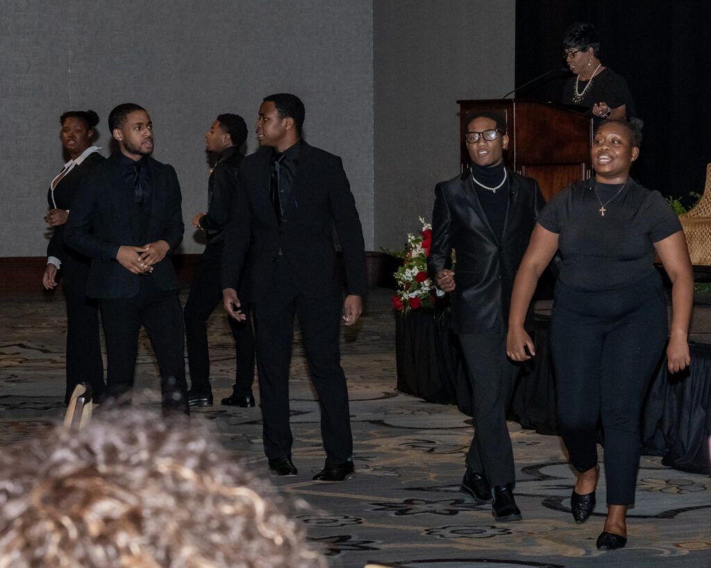 A group of people in black attire walks across a carpeted room. A person stands at a podium in the background. There are floral arrangements near the podium. The atmosphere seems formal.