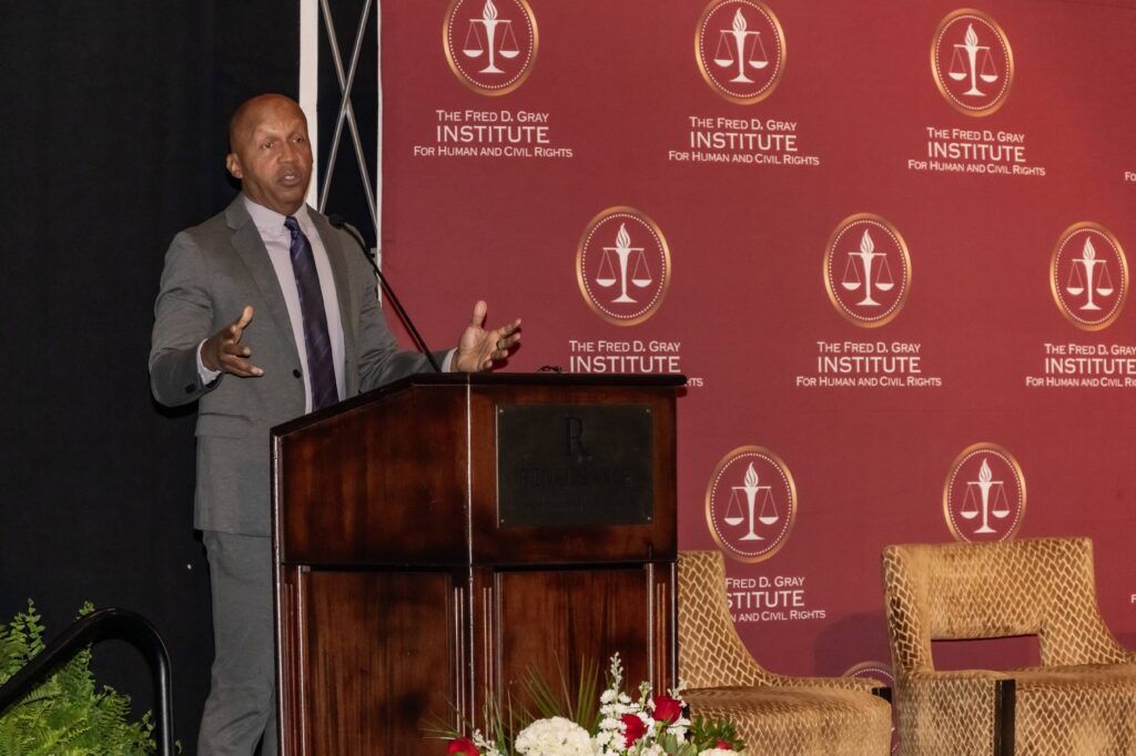A person speaks at a wooden podium with a microphone, gesturing with one hand. Behind them is a red backdrop with "The Fred D. Gray Institute for Human and Civil Rights" printed multiple times. Two wicker chairs are visible to the right.