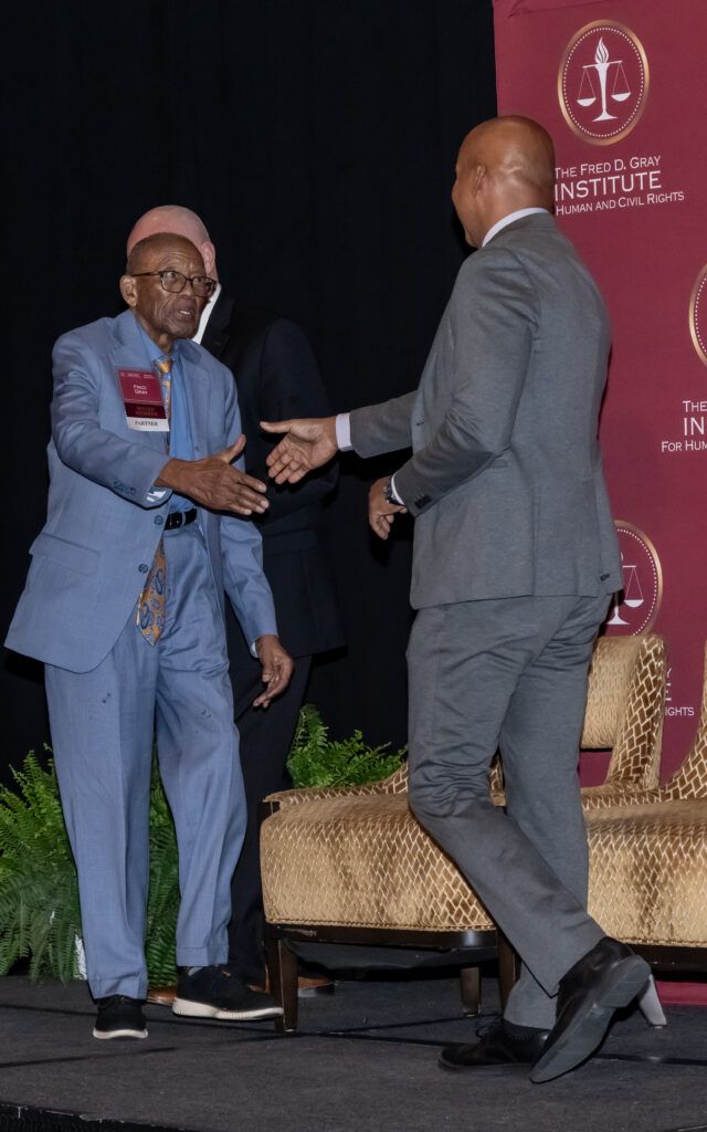 Two men shaking hands on a stage. The man on the left is wearing a light blue suit and the man on the right is in a gray suit. There is a red banner with a logo in the background, and the scene suggests a formal or professional event.