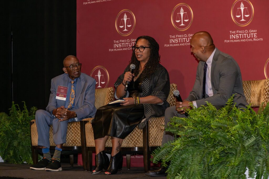 Three individuals are seated on a stage with a red backdrop that reads "The Fred D. Gray Institute for Human and Civil Rights." The center person is holding a microphone, and ferns are visible at the front of the stage.