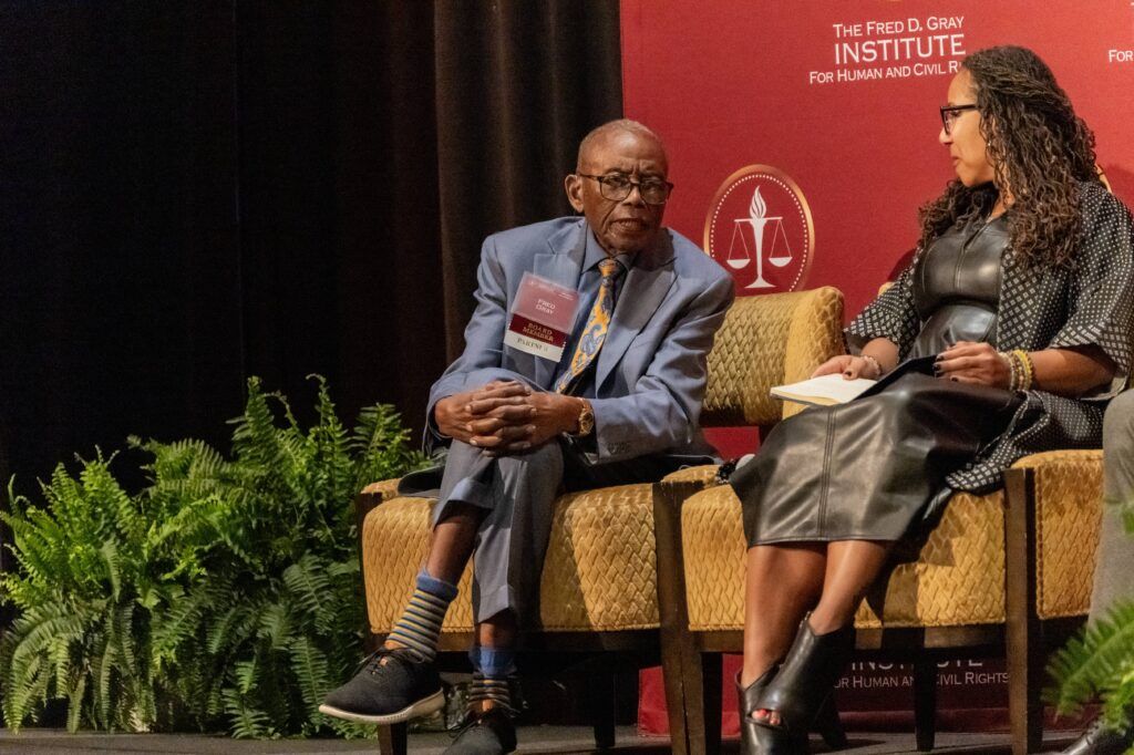 An elderly man in a suit sits on a stage next to a woman in a leather dress holding a book. They are part of a discussion at an event associated with the Fred D. Gray Institute. Ferns and a red backdrop are visible.