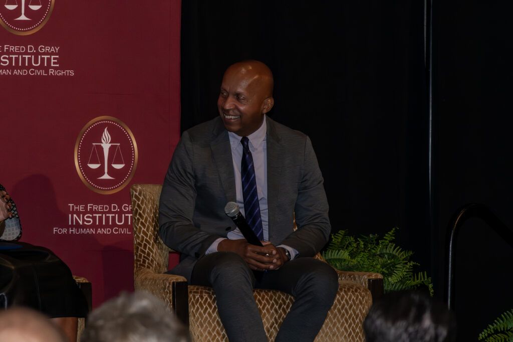 A person in a gray suit and striped tie sits on a wicker chair, smiling, with a maroon backdrop featuring logos and text that reads "The Fred D. Gray Institute for Human and Civil Rights.