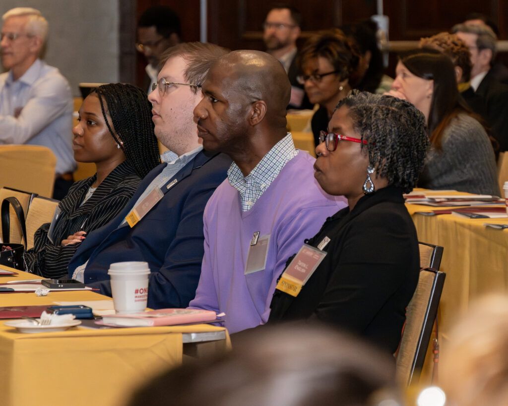 A group of people sitting in a conference room, attentively listening to a presentation. They are seated at tables covered with yellow tablecloths, with notepads and a coffee cup in front of them. The background includes more attendees and wood paneling.