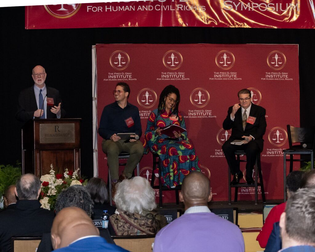 Three panelists are seated on a stage with red banners behind them at a symposium. A man stands at a podium on the left, speaking. Audience members are seated in the foreground.