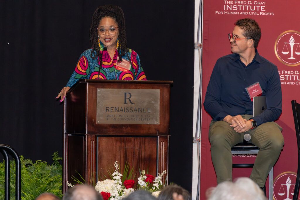 A woman stands at a podium with a colorful dress, speaking to an audience. A man sits to her right on a stage chair, facing her. The backdrop includes text about the Fred D. Gray Institute. Flowers are in front of the podium.