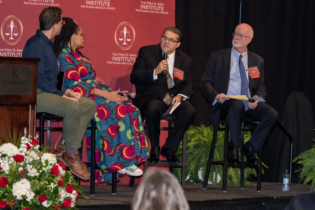 Four people are seated on a stage panel discussion at The Fred D. Gray Institute for Human and Civil Rights. One person is speaking into a microphone. A podium with flowers is visible on the left, and ferns are in front of a red backdrop.
