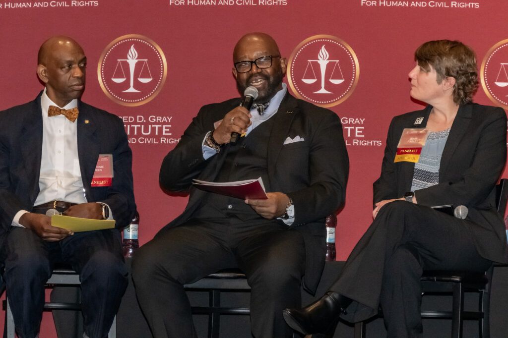Three individuals are seated on stage, participating in a panel discussion. The person in the center speaks into a microphone. They are dressed in formal attire against a backdrop with legal-themed graphics and text.