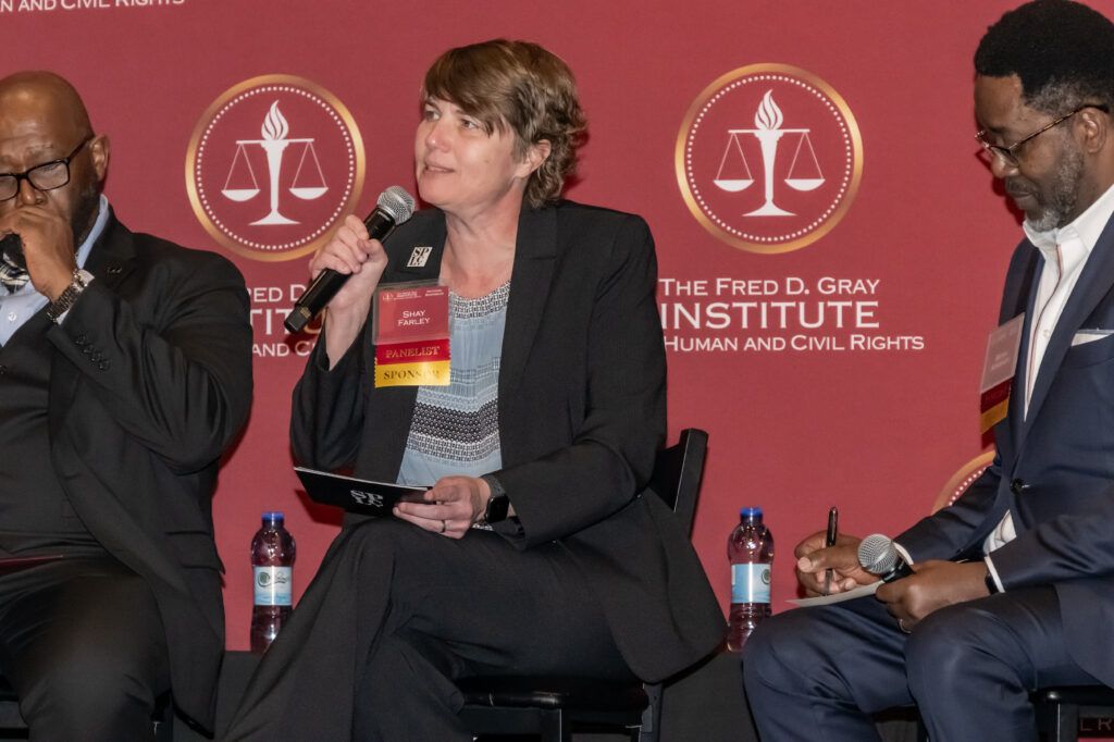 Three individuals are seated on a stage in front of a red backdrop with the Fred D. Gray Institute logo. The person in the center holds a microphone, speaking, while the others listen. Bottles of water are on the table in front of them.