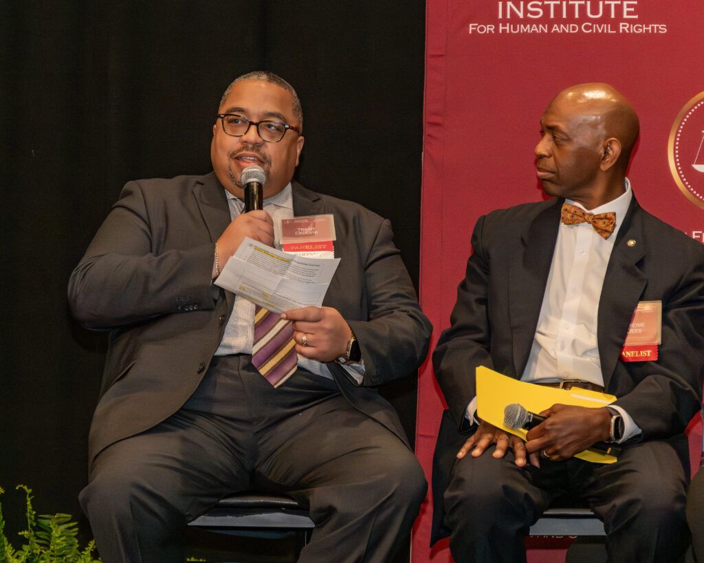 Two men in suits are sitting on a stage. The man on the left is speaking into a microphone while holding papers. The man on the right, wearing a bow tie, is holding a folder. A banner displaying "Institute for Human and Civil Rights" is in the background.