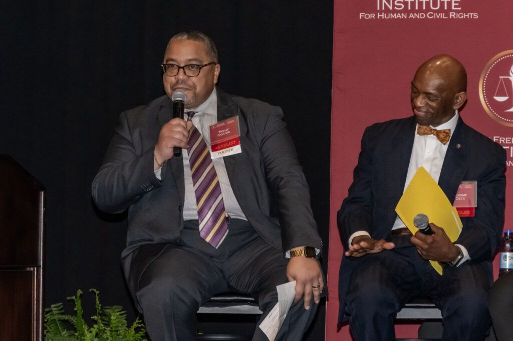 Two men in suits sit on stage. One speaks into a microphone and holds a paper. The other smiles, holding a yellow folder. A backdrop displays a logo with scales of justice. Both wear conference name tags.