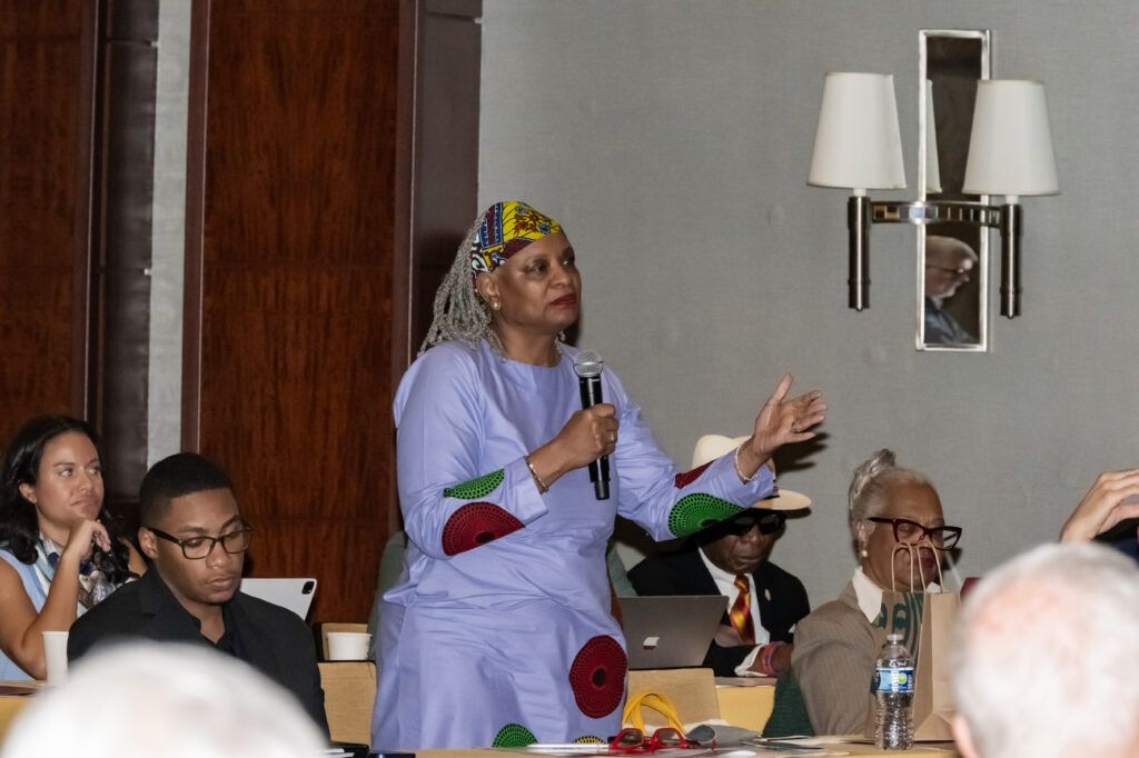 A person in a purple outfit speaks into a microphone while standing in a conference room. Others are seated nearby at a table with laptops and papers. Conference materials and a water bottle are visible on the table.