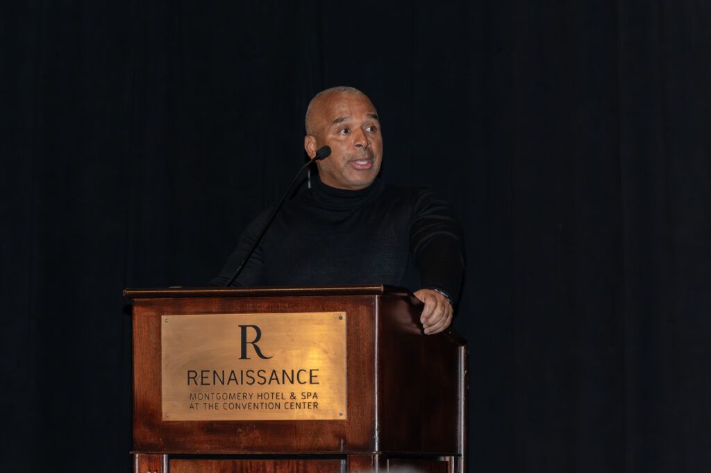 A person in a black turtleneck speaks at a podium with a Renaissance Montgomery Hotel & Spa sign. The background is dark.