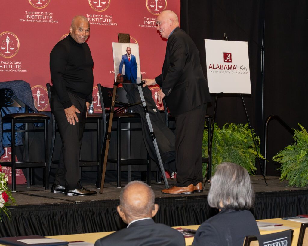 Two men stand on a stage at an event, one adjusting a photo on an easel. A podium and a sign for the Alabama Law School are in the background. Audience members are seated in the foreground.