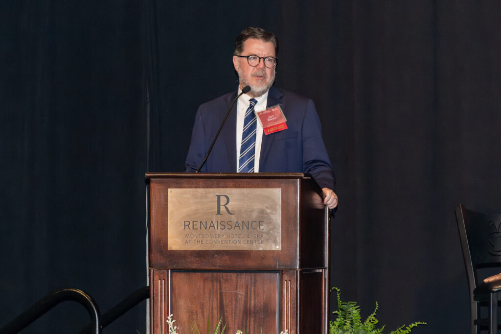 A man in a suit stands at a podium labeled "Renaissance" with a microphone. The background is a dark curtain, and there is a plant partially visible behind the podium. He appears to be speaking or giving a presentation.