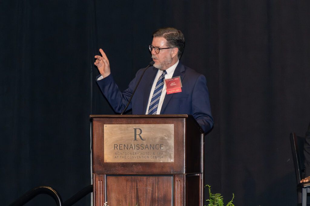 A man in a suit and tie speaks at a podium with the Renaissance logo on it. He appears to be giving a presentation or speech, gesturing with one hand raised. The background is dark and the setting seems formal.