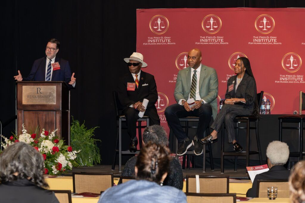 A man stands at a podium speaking to an audience. Three other individuals are seated on a panel to his right. They are in front of a red backdrop with logos and text. There is a floral arrangement near the podium, and people are seated in the foreground.