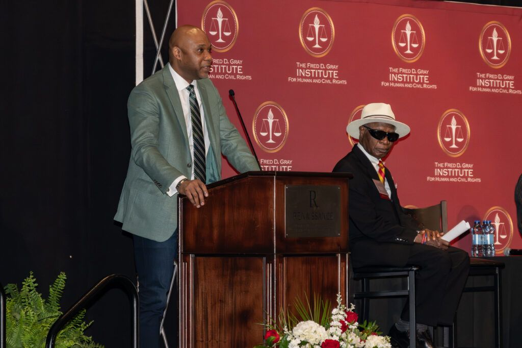 A man in a gray suit speaks at a podium on a stage, with another person in a hat and sunglasses seated nearby. The backdrop displays the logo and name of the Fred D. Gray Institute for Human and Civil Rights. Flowers are in the foreground.