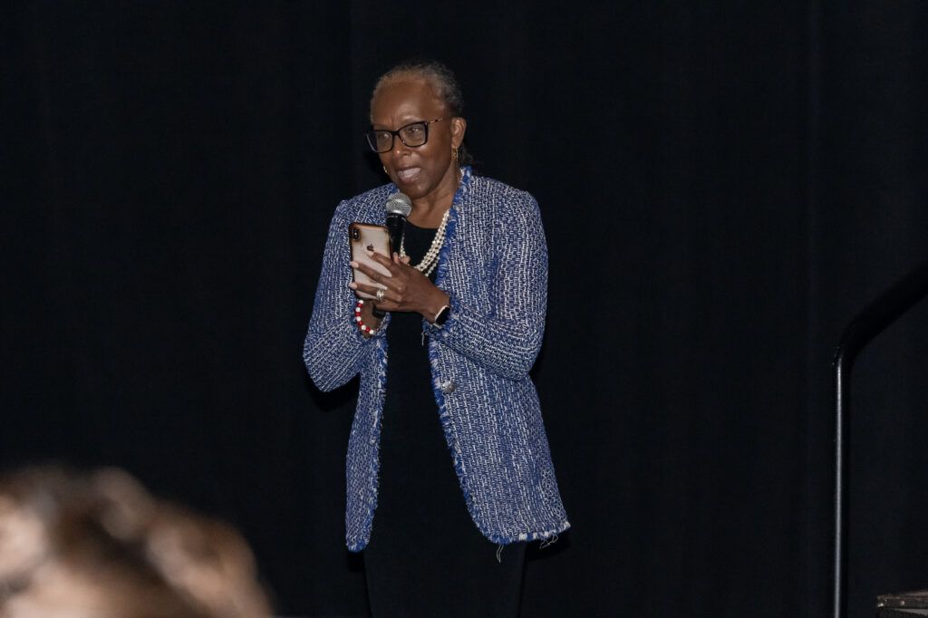 An individual with short hair and glasses is speaking into a microphone while holding a smartphone. They are wearing a blue patterned blazer over a black outfit. The background is dark.
